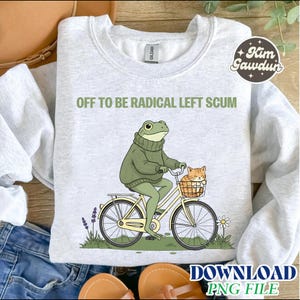 Könnte beinhalten: Hellgraues Sweatshirt mit dem Text "OFF TO BE RADICAL LEFT SCUM". Die Grafik zeigt einen Frosch in einem grünen Pullover auf einem gelben Fahrrad mit einer Katze im Korb. Eine Gänseblümchen und Lavendel sind zu sehen.
