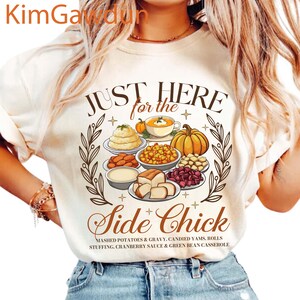 May include: Cream-colored t-shirt with a Thanksgiving-themed graphic. The design features a variety of dishes, including mashed potatoes, gravy, and a pumpkin. Text reads "JUST HERE for the Side Chick".