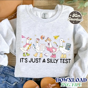 Op de afbeelding: Lichtgrijs sweatshirt met de tekst "IT'S JUST A SILLY TEST". Het ontwerp toont cartoon ganzen met accessoires zoals laarzen, hoeden en vlinderdassen, die items vasthouden zoals een potlood, testpapier en een rekenmachine.