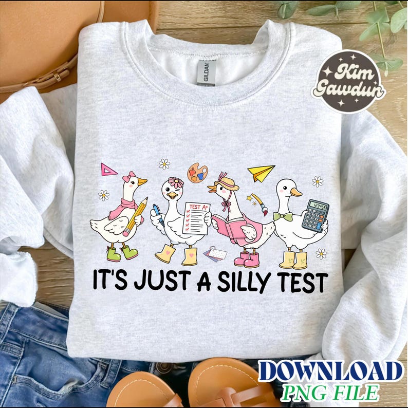 Op de afbeelding: Lichtgrijs sweatshirt met de tekst "IT'S JUST A SILLY TEST". Het ontwerp toont cartoon ganzen met accessoires zoals laarzen, hoeden en vlinderdassen, die items vasthouden zoals een potlood, testpapier en een rekenmachine.