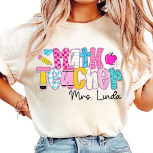 Könnte beinhalten: Cremefarbenes T-Shirt mit einem farbenfrohen "Math Teacher"-Aufdruck. Das Design umfasst einen Bleistift, ein Papierflugzeug, einen Apfel und eine Blume. Der Name "Mrs. Linda" ist darunter gedruckt.
