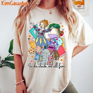 May include: A white t-shirt with a colorful graphic print of a teacher and students. The graphic includes a teacher with a microphone, a fish, a book, a pencil, a cookie, a mouse, an elephant, a pig, and a seed. The text "In da clerk, we all fam" is printed below the graphic.