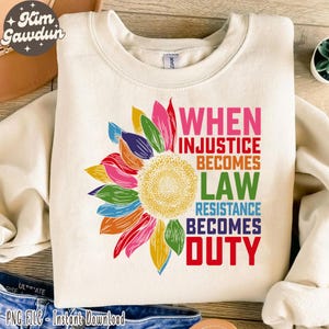 Pode incluir: Uma camisa de moletom branca com um gráfico de girassol colorido e o texto "WHEN INJUSTICE BECOMES LAW RESISTANCE BECOMES DUTY".