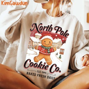 May include: Cream sweatshirt with a gingerbread man in a Santa hat and scarf, and the text "North Pole Cookie Co." and "Baked Fresh Daily." The design includes snowy houses and a red background.