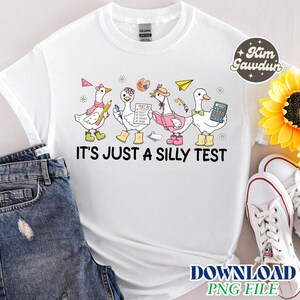 Op de afbeelding: Wit T-shirt met een grillig ontwerp van cartoon ganzen, elk met schoolspullen. De tekst "IT'S JUST A SILLY TEST" staat eronder. Een zonnebloem en een spijkerbroek zijn op de achtergrond.