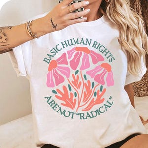 May include: White t-shirt with a graphic design of pink flowers and green leaves. The text "BASIC HUMAN RIGHTS ARE NOT "RADICAL"" is printed in green on the shirt.