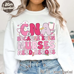 Puede incluir: Sudadera blanca con un diseño gráfico rosa y blanco que dice "CNA because even nurses need heroes". El gráfico incluye un estetoscopio, una venda, un frasco de pastillas, un corazón y un botiquín de primeros auxilios.