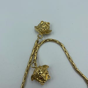 May include: Gold-toned necklace featuring two Medusa head pendants. The chain has a delicate, linked design. The pendants and chain are a bright, shiny gold color. The necklace is displayed against a plain white background.