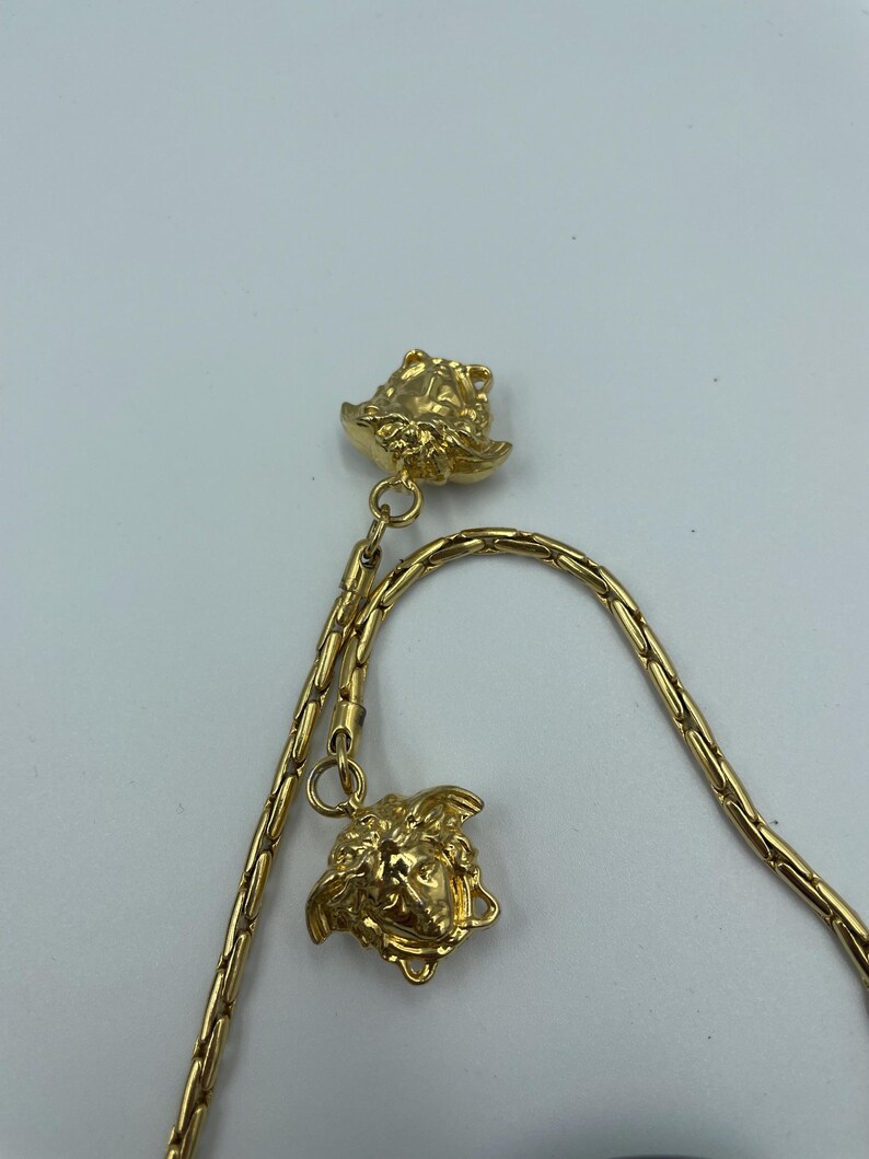 May include: Gold-toned necklace featuring two Medusa head pendants. The chain has a delicate, linked design. The pendants and chain are a bright, shiny gold color. The necklace is displayed against a plain white background.
