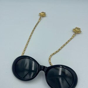 May include: Black oval sunglasses with gold-colored chain temples. The chain temples are attached to the frames with a gold-colored rose detail. The sunglasses have a glossy finish and are set against a white background.
