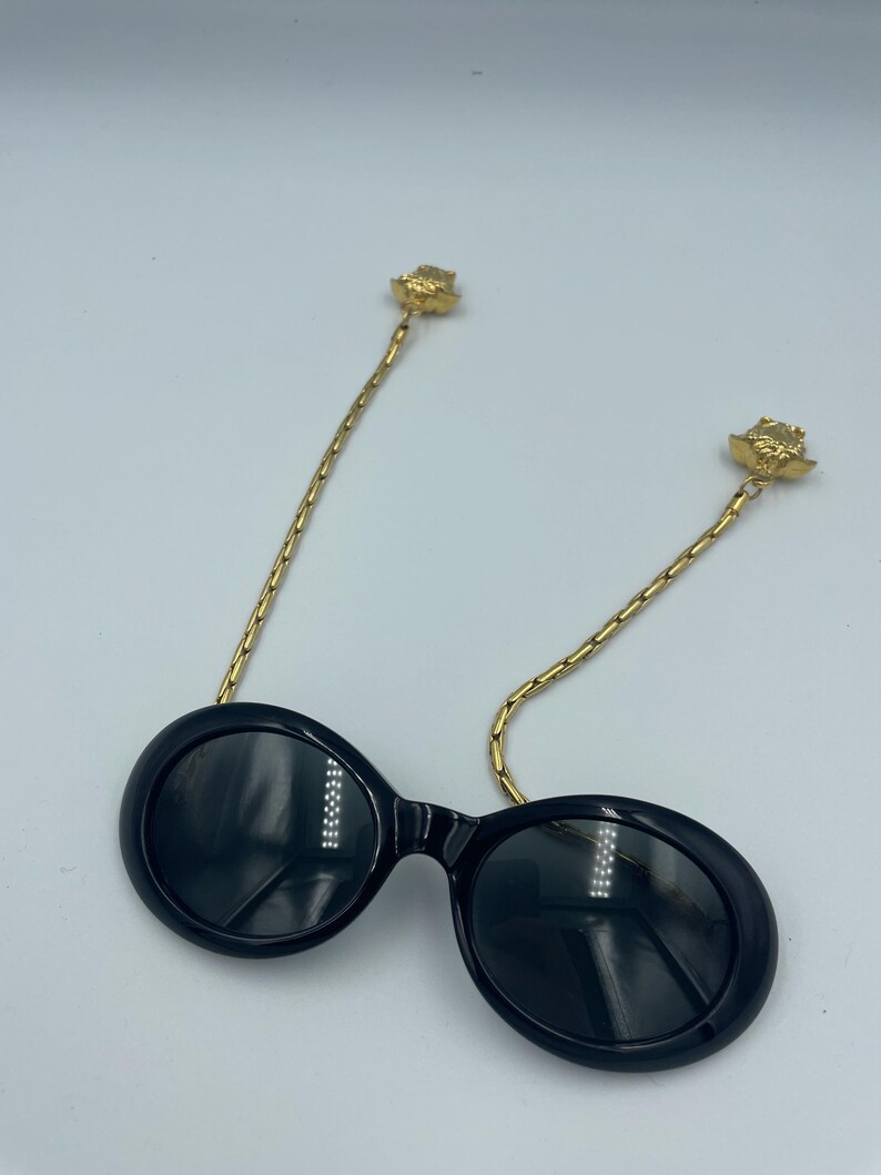 May include: Black oval sunglasses with gold-colored chain temples. The chain temples are attached to the frames with a gold-colored rose detail. The sunglasses have a glossy finish and are set against a white background.