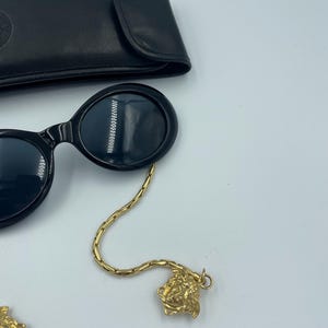 May include: Black oval sunglasses with a gold chain and Medusa head charm. The sunglasses are next to a black leather case. The chain is attached to the sunglasses and has a gold Medusa head charm at the end.