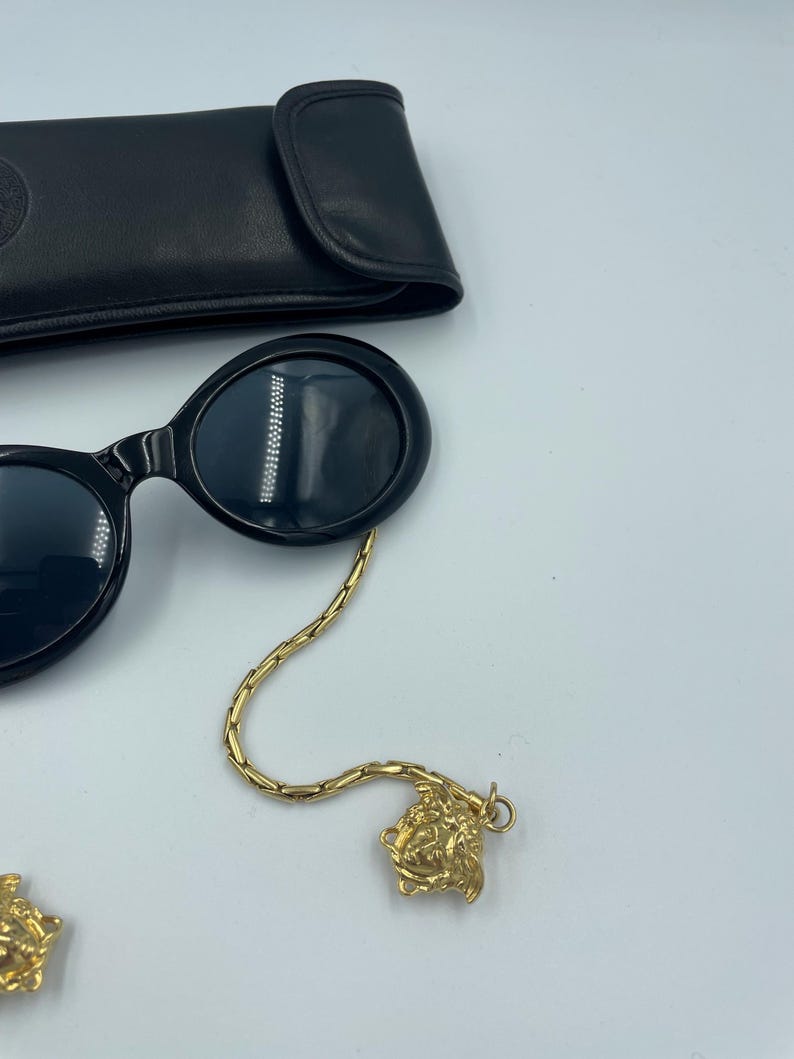 May include: Black oval sunglasses with a gold chain and Medusa head charm. The sunglasses are next to a black leather case. The chain is attached to the sunglasses and has a gold Medusa head charm at the end.