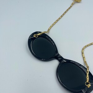 May include: Black oval sunglasses with a gold chain and a decorative gold Medusa head. The sunglasses have the text "GIANNI VERSACE MADE IN ITALY" and "MOD 403M COL 852".