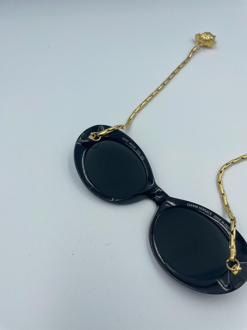 May include: Black oval sunglasses with a gold chain and a decorative gold Medusa head. The sunglasses have the text "GIANNI VERSACE MADE IN ITALY" and "MOD 403M COL 852".