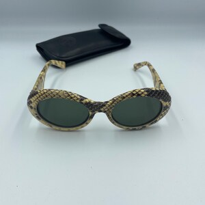 May include: Oval-shaped sunglasses with a snakeskin pattern on the frames and dark green lenses. A black leather case is behind the sunglasses. The sunglasses have a vintage aesthetic.