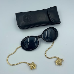 May include: Black oval sunglasses with gold chain and Medusa head accents, and a black leather case. The sunglasses have dark lenses and a sleek, modern design. The gold chain adds a touch of luxury.