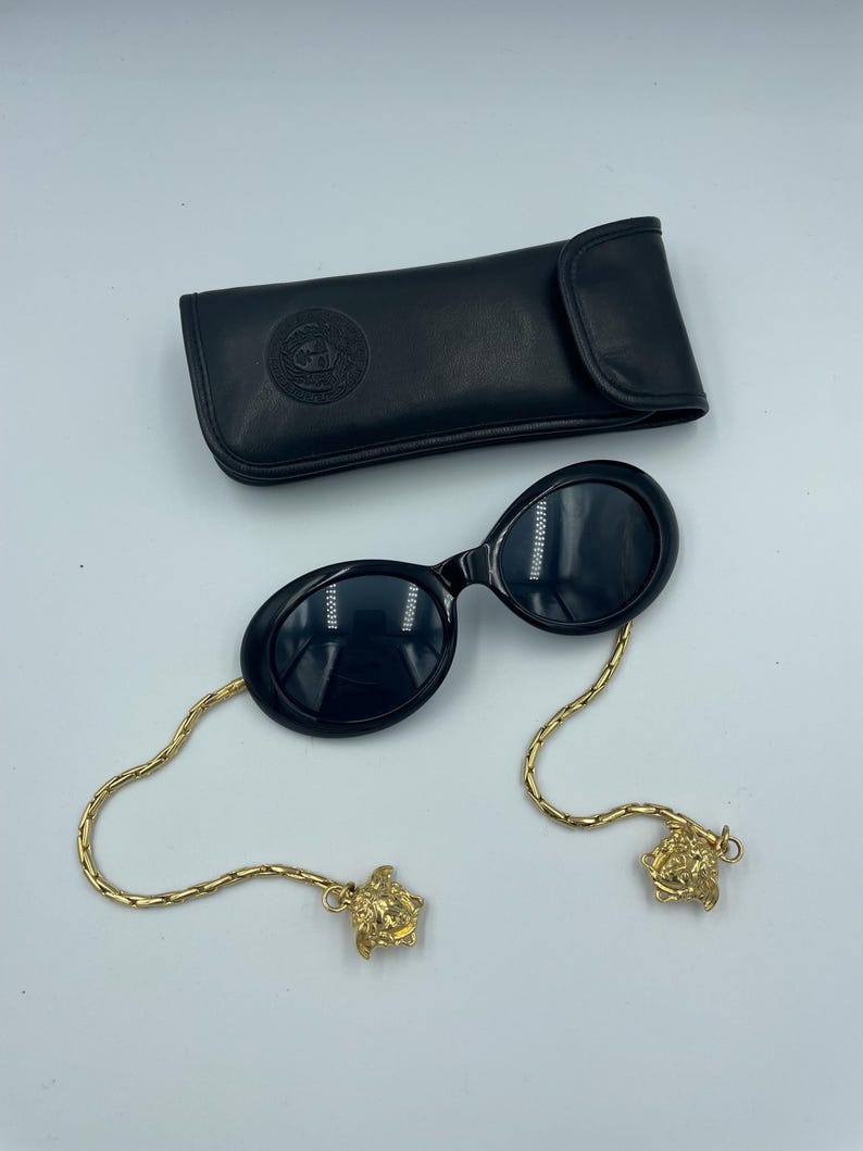 May include: Black oval sunglasses with gold chain and Medusa head accents, and a black leather case. The sunglasses have dark lenses and a sleek, modern design. The gold chain adds a touch of luxury.