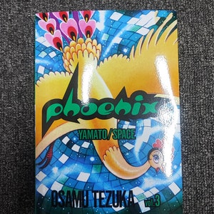 May include: A green and black "phoenix" logo with the words "YAMATO/SPACE" and "OSAMU TEZUKA" below it. The logo is on a blue and white background with a yellow bird with a red beak and a pink and purple tail.