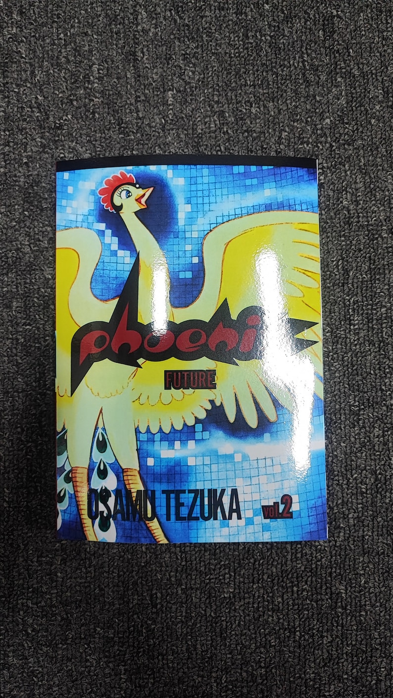 May include: A comic book cover featuring a yellow bird with red and black text that reads "Phoenix Future" and "Osamu Tezuka Vol. 2". The background is a blue and white mosaic pattern.