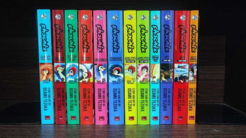 May include: A collection of 12 colorful graphic novels titled "Phoonix" by Osamu Tezuka. The books are arranged in order from volume 1 to volume 12. Each volume has a different cover design and title, including "Dawn", "Forever", "20th Century", "Karma", "Resurrection", "Nostalgia", "Civil War Part One", "Civil War Part Two", "Change to Survive", "Sun Part One", "Sun Part Two", and "Early Works".