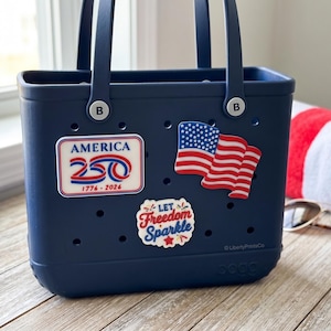 May include: A navy blue tote bag with two handles, adorned with patriotic stickers including an American flag and text that reads "America 250 1776-2026" and "Let Freedom Sparkle". A red and white striped towel and sunglasses are in the background.