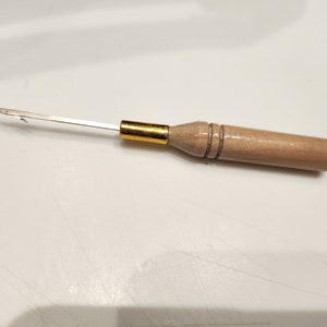 May include: A wooden crochet hook with a gold metal tip and a silver metal hook. The hook is about 6 inches long.