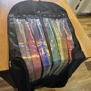 Puede incluir: Una funda de ropa negra con cremallera, que contiene múltiples extensiones de cabello de colores en fundas de plástico transparente. Las extensiones son de varios colores, incluyendo rosa, azul, verde y rojo, con detalles de purpurina. La bolsa está abierta sobre una superficie de madera.