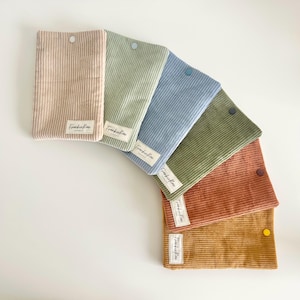 May include: A selection of corduroy pouches in shades like pale pink, green, and blue. Each pouch features a button fastening and a small white label with the text "Frankie Rae Handmade". The pouches are arranged in a fan shape, showcasing their colours.