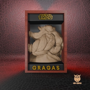 Puede incluir: Una caja de madera marrón con un interior negro y una inscripción dorada que dice "League of Legends". Dentro de la caja hay una figura beige de Gragas, un personaje del juego League of Legends. La figura está en una pose de lucha con una gran barba y un barril en la mano. La caja tiene una inscripción dorada que dice "Gragas".