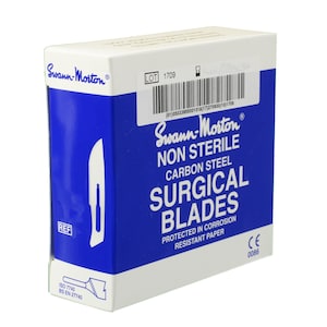 May include: A box of Swann-Morton surgical blades. The box is white and blue, with the brand name and product information printed on the front. The text on the box includes "NON STERILE", "CARBON STEEL", and "SURGICAL BLADES".