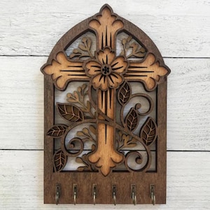 May include: Wooden key holder with a cross and floral design. The cross is brown with a lighter brown center. The floral design is in a lighter brown color. The key holder has six hooks.
