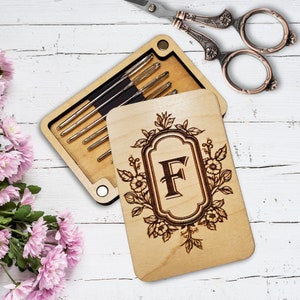 May include: A wooden box with a floral design and the letter "F" engraved on the lid. The box is open and contains a set of needles.