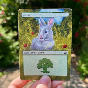 May include: A custom-painted "Forest" Magic: The Gathering card featuring a watercolor-style illustration of a rabbit in a field. The card has the text "Forest" at the top and "Basic Land - Forest" at the bottom, with a tree symbol.