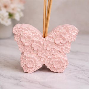 Può includere: Un diffusore rosa a forma di farfalla con un design floreale. Il diffusore ha una superficie strutturata e contiene diversi bastoncini di legno. Lo sfondo è una superficie simile al marmo.