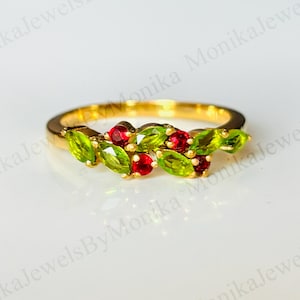 Puede incluir: Un anillo de oro con un grupo de piedras preciosas verdes y rojas. El diseño del anillo incorpora piedras verdes con corte marquesa y piedras rojas redondas, creando un patrón floral o similar a una baya. El anillo está sobre un fondo blanco.