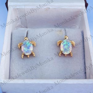 May include: A pair of gold turtle earrings with iridescent opal stones. The earrings are in a white box with the text "Monika Jewels By Monika" on the box.