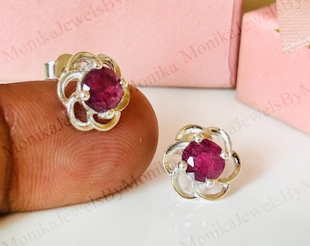 Peut inclure: Une paire de boucles d'oreilles à clous en argent avec un motif floral. Chaque boucle d'oreille présente une pierre précieuse rubis ovale vibrante au centre, entourée de détails argentés délicats en forme de pétales. Les boucles d'oreilles sont présentées sur une surface rose clair.
