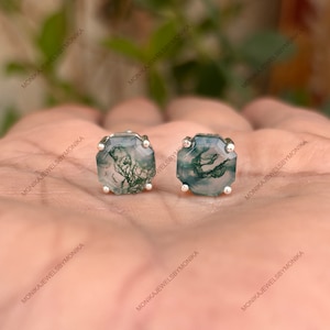 May include: A pair of square-cut moss agate stud earrings. The earrings feature a silver setting and showcase the natural green and white patterns within the stone. The earrings are displayed on a neutral background.