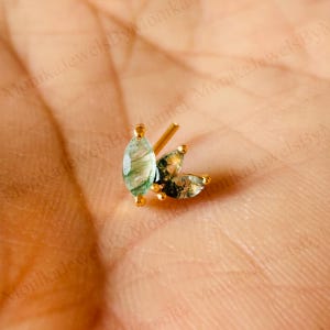 Puede incluir: Un pendiente de botón dorado con tres piedras preciosas en forma de marquesa, verdes y blancas, con un diseño similar al musgo. El poste del pendiente es visible. El pendiente está sobre una superficie marrón claro.