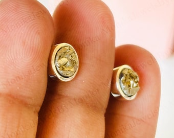Op de afbeelding: Een paar zilveren oorstekers met elk een goudkleurige, getextureerde steen. De oorbellen zijn rond en hebben een eenvoudig ontwerp. De oorbellen worden tussen de vingers gehouden.