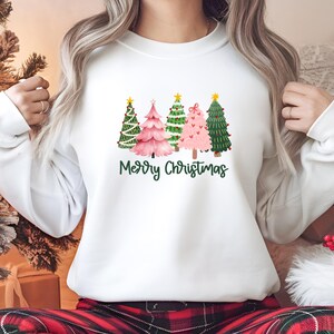 Könnte beinhalten: Weißes Sweatshirt mit einem Weihnachtsdesign. Es zeigt fünf Weihnachtsbäume in Grün und Rosa, jeder mit einem goldenen Stern. Die Worte "Merry Christmas" stehen in grüner Schrift darunter. Ein festliches Kleidungsstück zum Thema Feiertage.