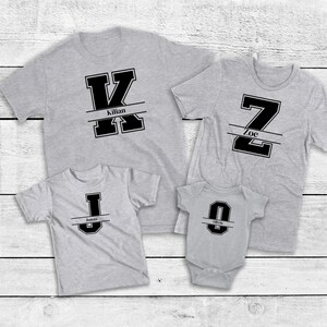 Könnte beinhalten: Vier graue T-Shirts und ein Body mit schwarzen Blockbuchstaben-Designs. Die Shirts zeigen die Buchstaben K, Z und J, jeweils mit einem Namen darunter. Der Body hat den Buchstaben O mit einem Namen darunter. Alles auf weißem Holzgrund.