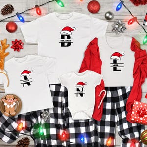 Könnte beinhalten: Weiße Familien-T-Shirts mit Weihnachtsmotiven und ein Strampler mit Weihnachtsmützen und personalisierten Initialen. Die Shirts sind mit schwarz-weiß karierten Schlafanzughosen kombiniert. Festliche Dekorationen umfassen Ornamente, Lichter und einen Lebkuchenmann.