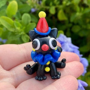 May include: A small, black figurine of a cat dressed as a clown. The cat has large white eyes, a red nose, and a blue ruffled collar. It wears a red and yellow clown hat. The figurine is held in a person's hand.