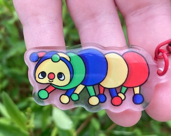 Peut inclure: Un porte-clés chenille coloré avec un anneau en métal rouge. La chenille a un design de dessin animé avec des segments verts, bleus, jaunes et rouges. Le porte-clés est en acrylique transparent.