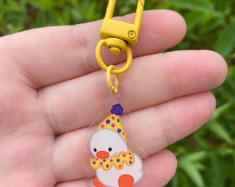 Peut inclure: Un porte-clés jaune avec un charm en forme de canard de dessin animé. Le canard est blanc avec des pieds oranges, un chapeau à pois jaunes et violets et un col à pois jaunes. Le porte-clés a un fermoir et un anneau jaunes.