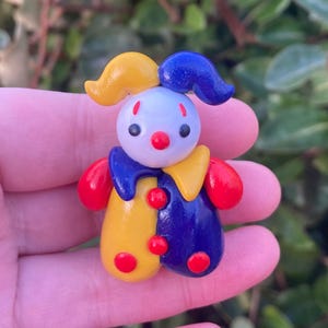 May include: A colorful, handcrafted clown figurine. The clown features a white head, red nose, and black eyes. It wears a blue and yellow outfit with red accents. The clown has a yellow hat and blue hair.
