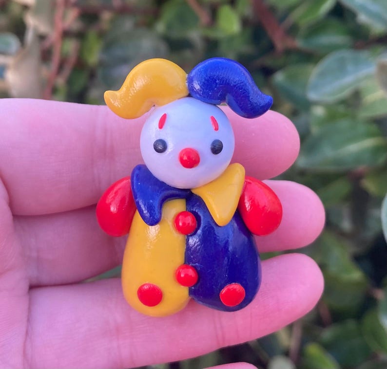 May include: A colorful, handcrafted clown figurine. The clown features a white head, red nose, and black eyes. It wears a blue and yellow outfit with red accents. The clown has a yellow hat and blue hair.