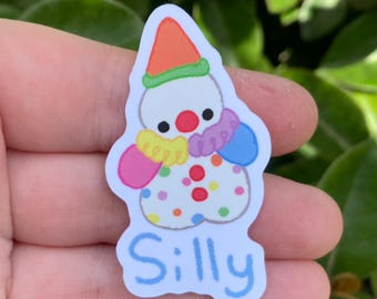 May include: A colorful sticker featuring a cartoon clown with a white body, orange hat, and multi-colored accents. The word "Silly" is written in blue script at the bottom. The clown has a red nose and polka dots.